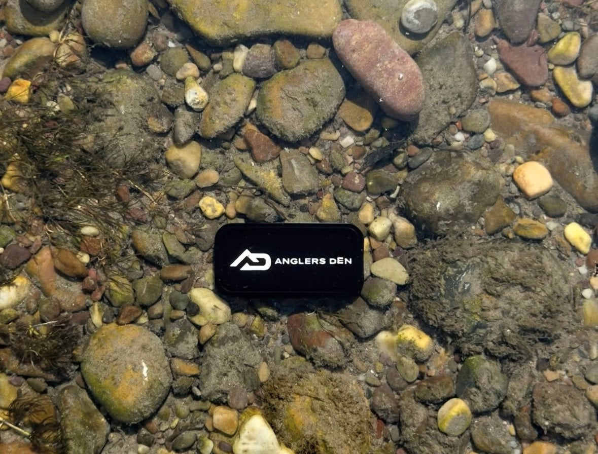 A black rectangular electronic device with the white logo and text 'ANGLERS DEN' resting on a bed of wet river stones and pebbles in shallow, clear water.
